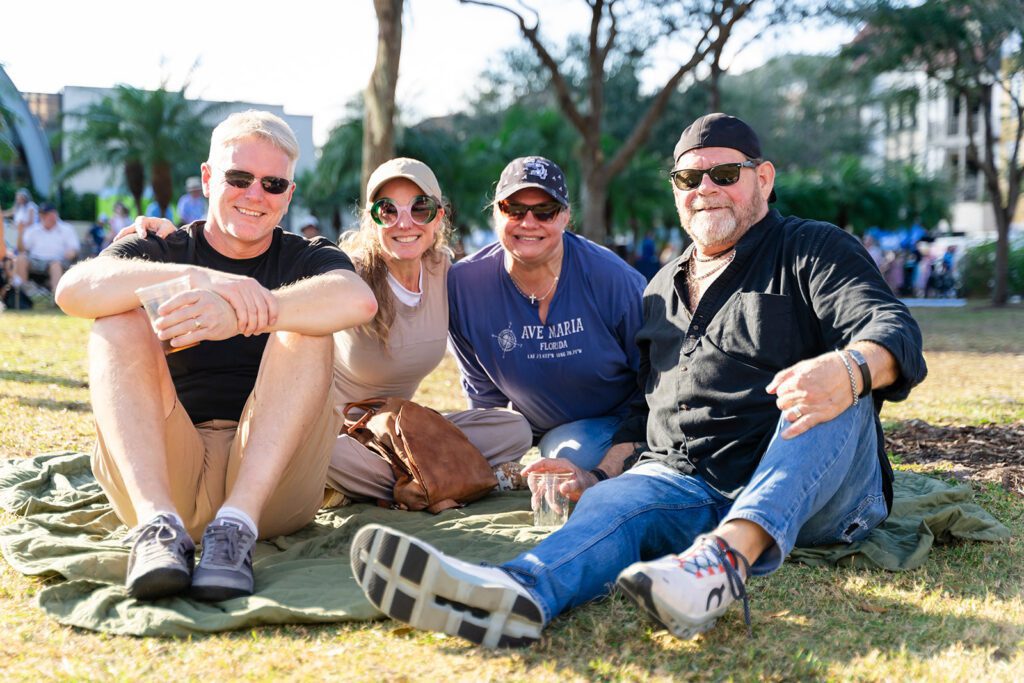 Group of four adult friends sit on the grass, witnessing the festivities of the 2025 Taste of Ave